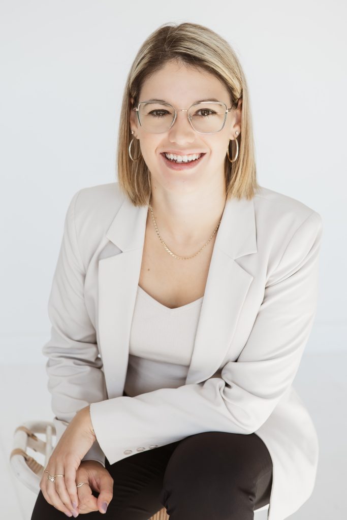 A woman wearing glasses, a white blouse, and a tan corduroy blazer sits at a table with her hands clasped during a professional meeting